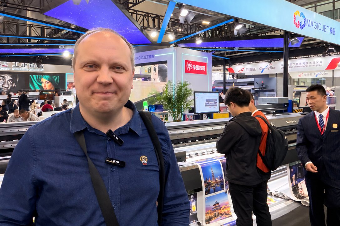 Сергей Белокуров, автор канала Inkjet Всемогущий, на стенде MAGICJET: позади широкоформатный рулонный принтер печатает городские пейзажи